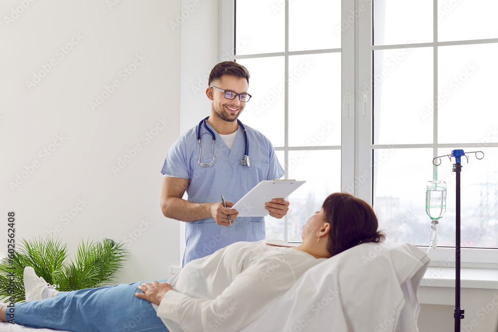 A smiling healthcare professional reading information from a clipboard to a patient lying on a hospital bed.