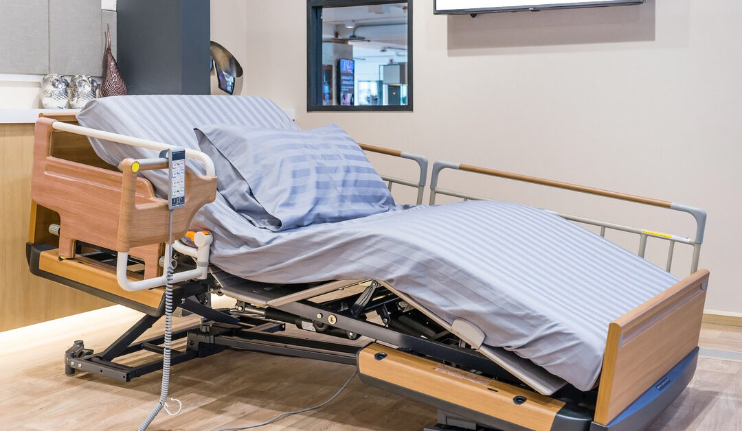 A hospital bed sits empty in a clean patient room. The mattress curves to elevate the head section and lower the foot section.