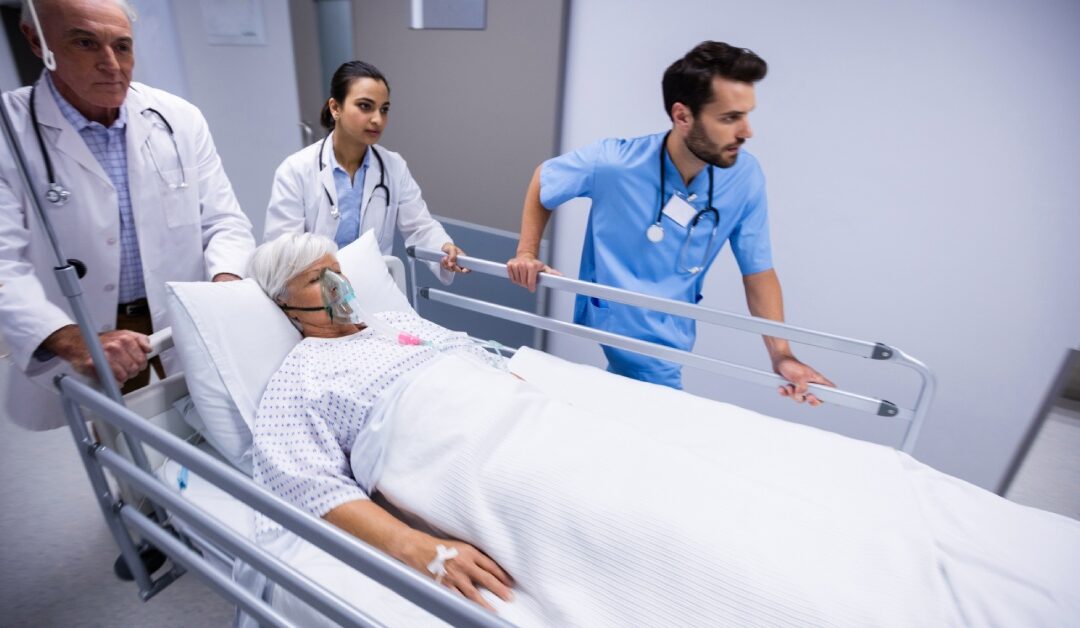 Three healthcare professionals pushing a patient lying on a stretcher down a hospital hallway. The patient wears an oxygen mask.