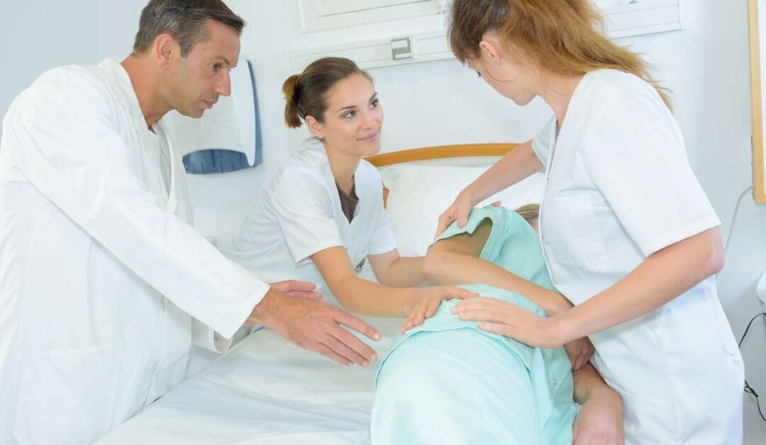 A doctor in a white coat and two other healthcare workers gently repositioning a patient onto their side in a hospital bed.