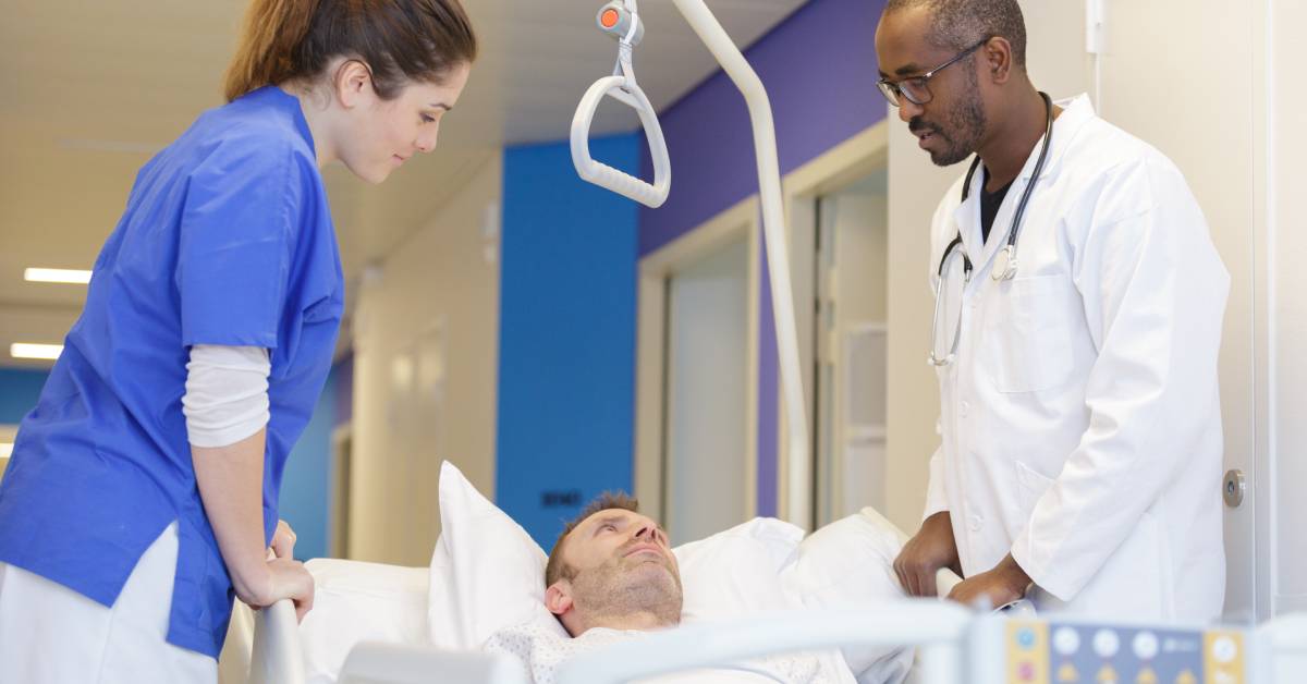 Two healthcare workers transporting a patient in a hospital bed down a hallway. They are looking at the patient's face.