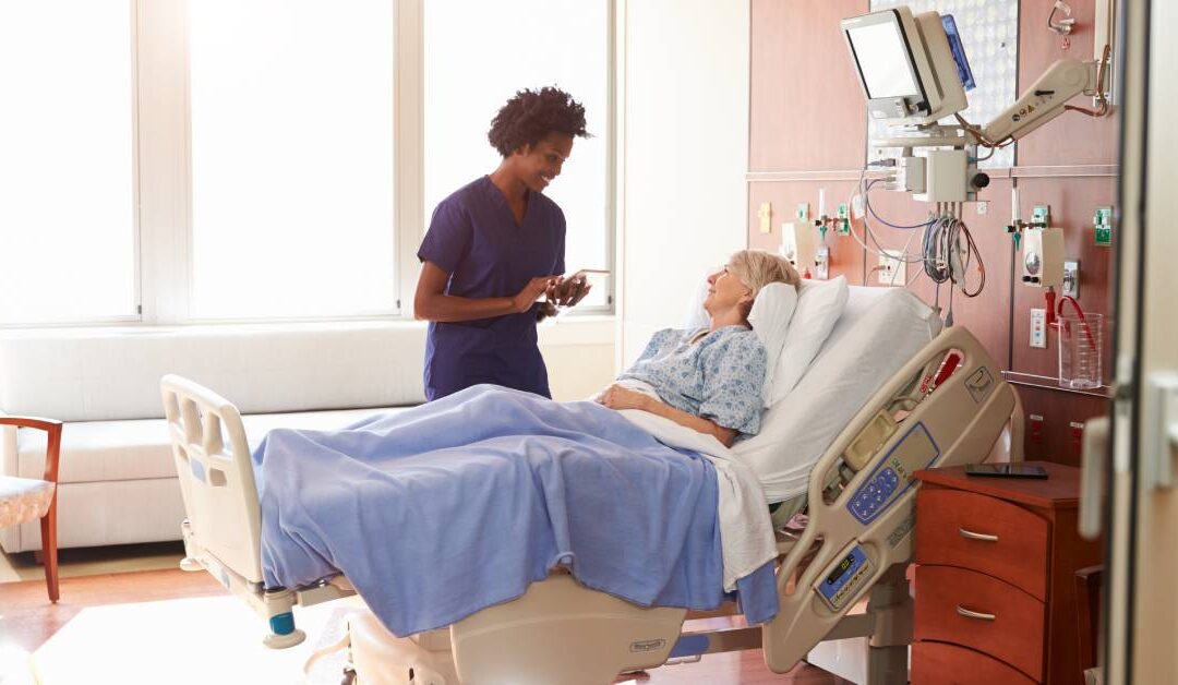 A nurse holding a digital tablet while talking to a patient who is in a hospital bed. The headrest is raised at a 45-degree angle.