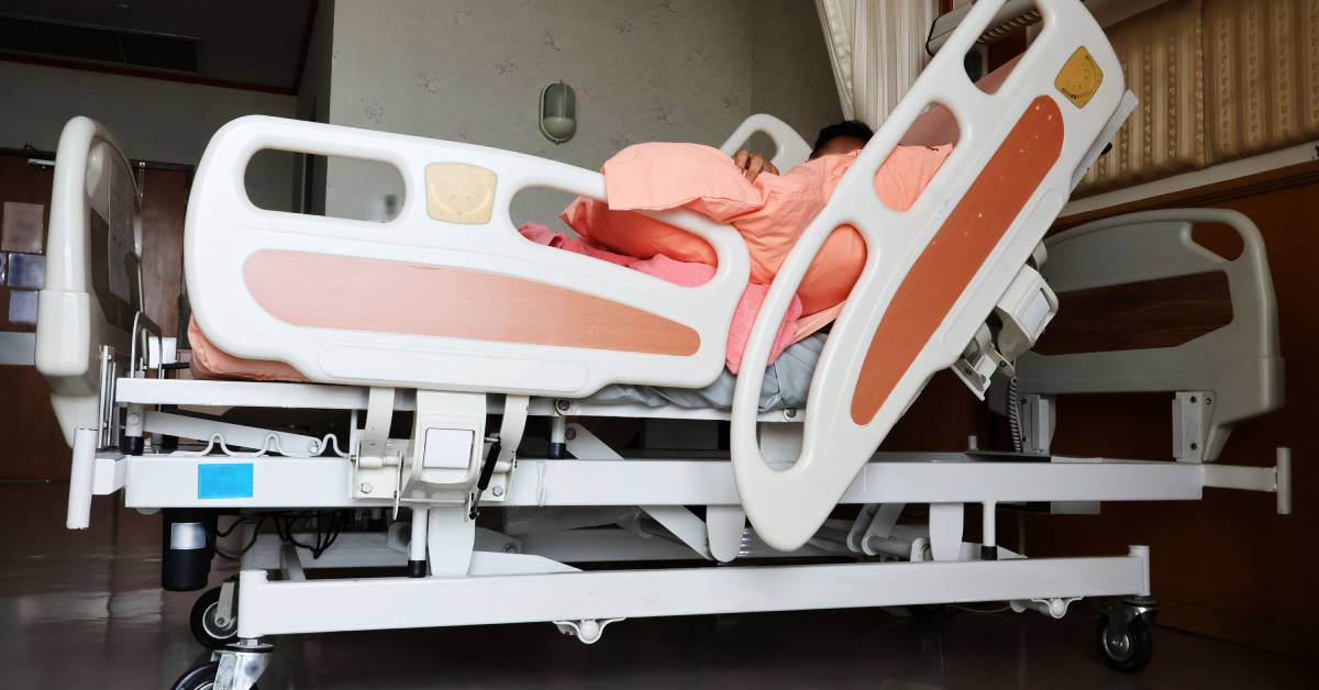 A side view of a hospital bed with a raised headrest and raised side rails. Pink bedding is pressed against the side rail.