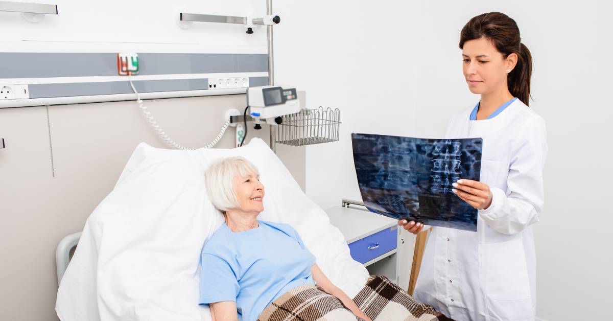 A smiling doctor in a white lab coat stands beside a patient lying in a hospital bed. The doctor holds a radiograph.