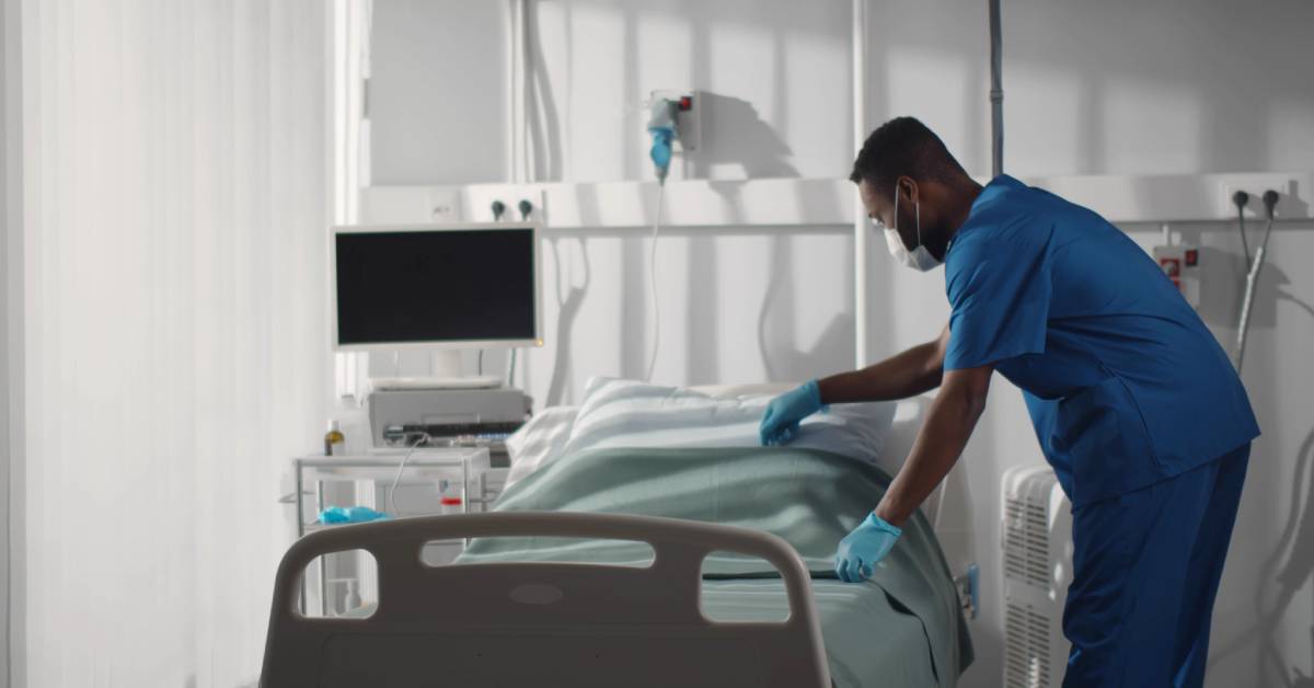 A medical professional wearing scrubs, a mask, and gloves adjusts the blanket on a hospital bed in an empty patient room.