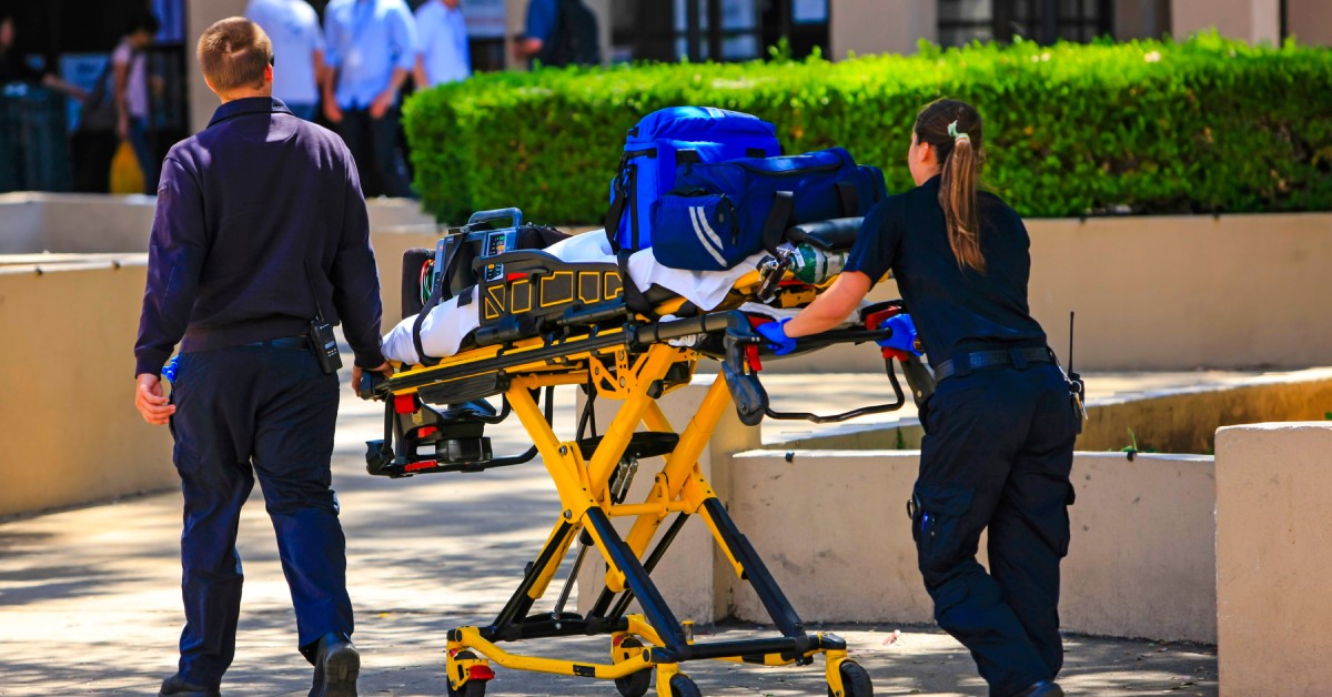 Two paramedics push a yellow stretcher on a sidewalk by a building. Bags of medical equipment are on the stretcher mattress.