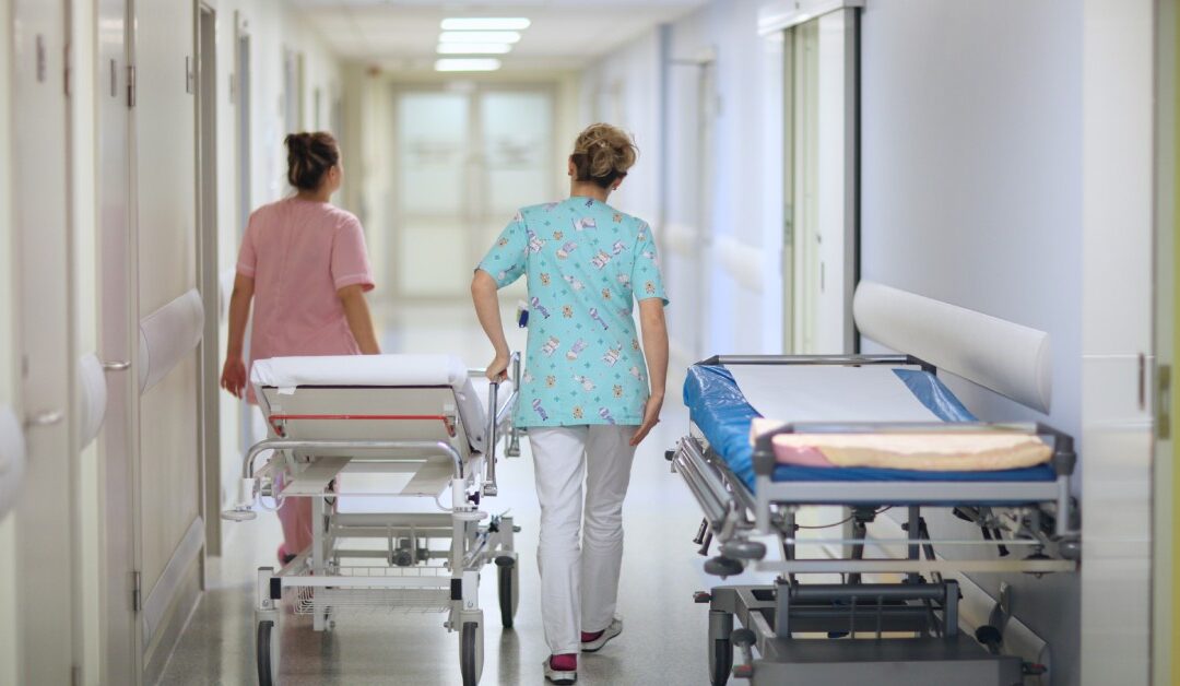 Two hospital workers wearing scrubs push a stretcher in a hospital corridor. Another stretcher is parked against a wall.