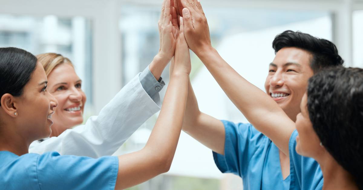 Four smiling healthcare workers standing in a circle. They each put one hand in the center of the circle for a group high-five.