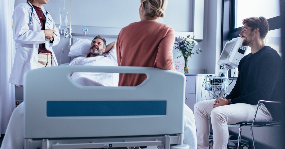 A patient and visitors talking to a doctor. The patient is in a hospital bed and one visitor is sitting at the foot of the bed.