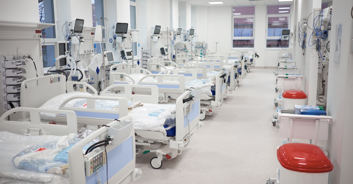 A row of empty hospital beds set against a wall in a brightly lit hospital room. Items in sealed bags are on top of each bed.