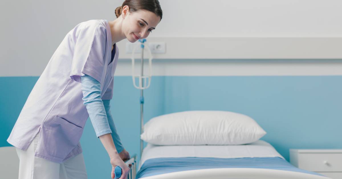 A person in scrubs adjusting the rail on one side of a hospital bed. A blue blanket and a thick pillow are on the bed.