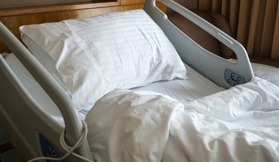 Natural light illuminating the white bedding and pillow on a hospital bed. Bed rails are raised on either side of the pillow.