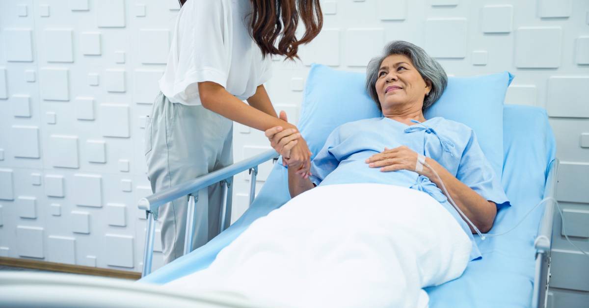 An older adult lying in a hospital bed with the side rails up. She is smiling and holding the hand of a person next to her.