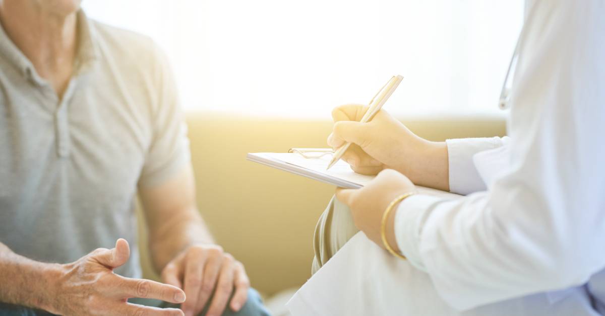 A patient seated on a couch is talking and gesticulating. The doctor across from the patient is taking notes on a clipboard.