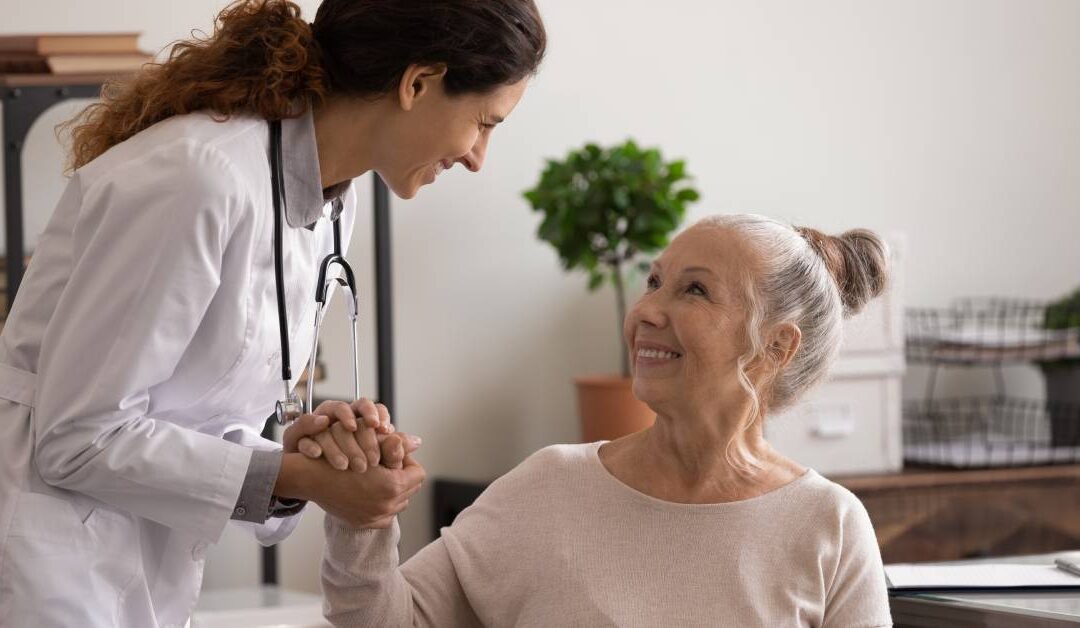 A doctor wearing a white lab coat stands next to a seated patient and clasps her hand. The patient is smiling.