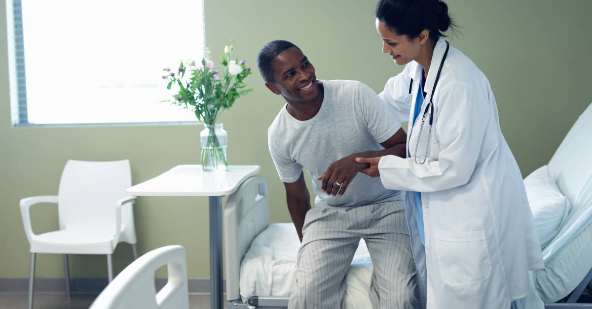 A doctor helps a smiling patient stand up from a hospital bed. The head of the bed is raised about 60 degrees.