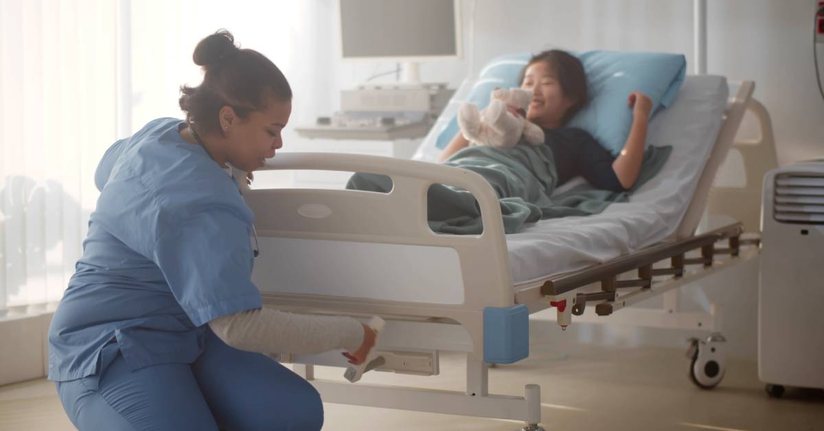 A healthcare worker kneels and adjusts the footboard panel on a hospital bed. The patient lies in bed and holds a soft toy.