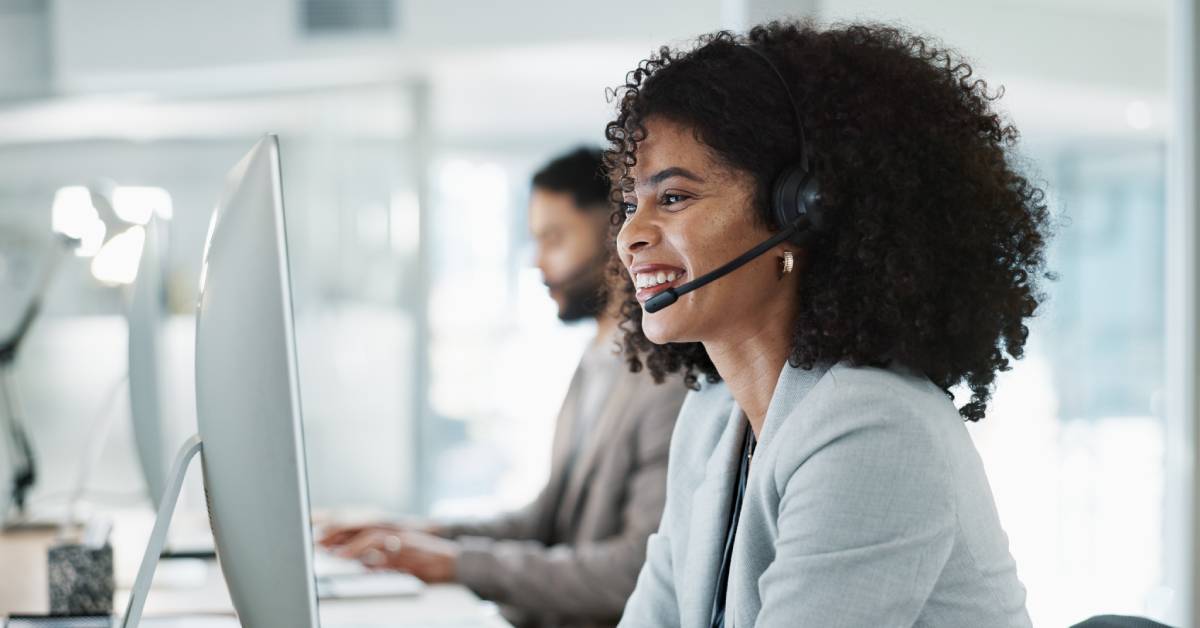 A woman wearing a headset smiles and looks at her computer screen. She is dressed professionally and sits next to a coworker.